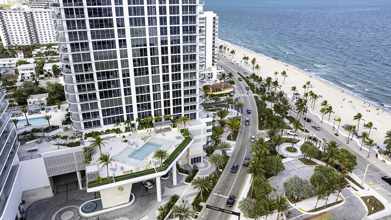 Aerial drone view of Selene Oceanfront Residences arrival area in Fort Lauderdale, showing the sleek 26-story white towers, elevated resort-style pools, palm-lined entrance, and direct Atlantic Ocean beachfront along A1A.