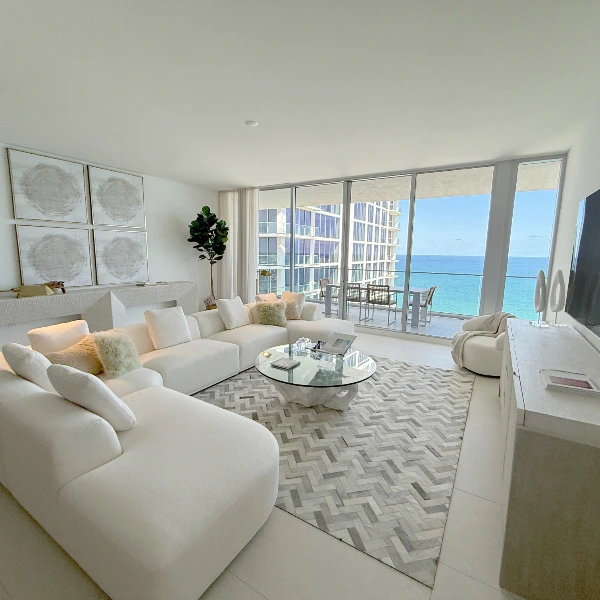 Bright modern living room with curved white sofas, glass coffee table, and balcony overlooking the ocean.